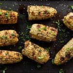Grilled corn on the cob garnished with lime wedges, sesame seeds and green herbs on a baking sheet with glasses of beer