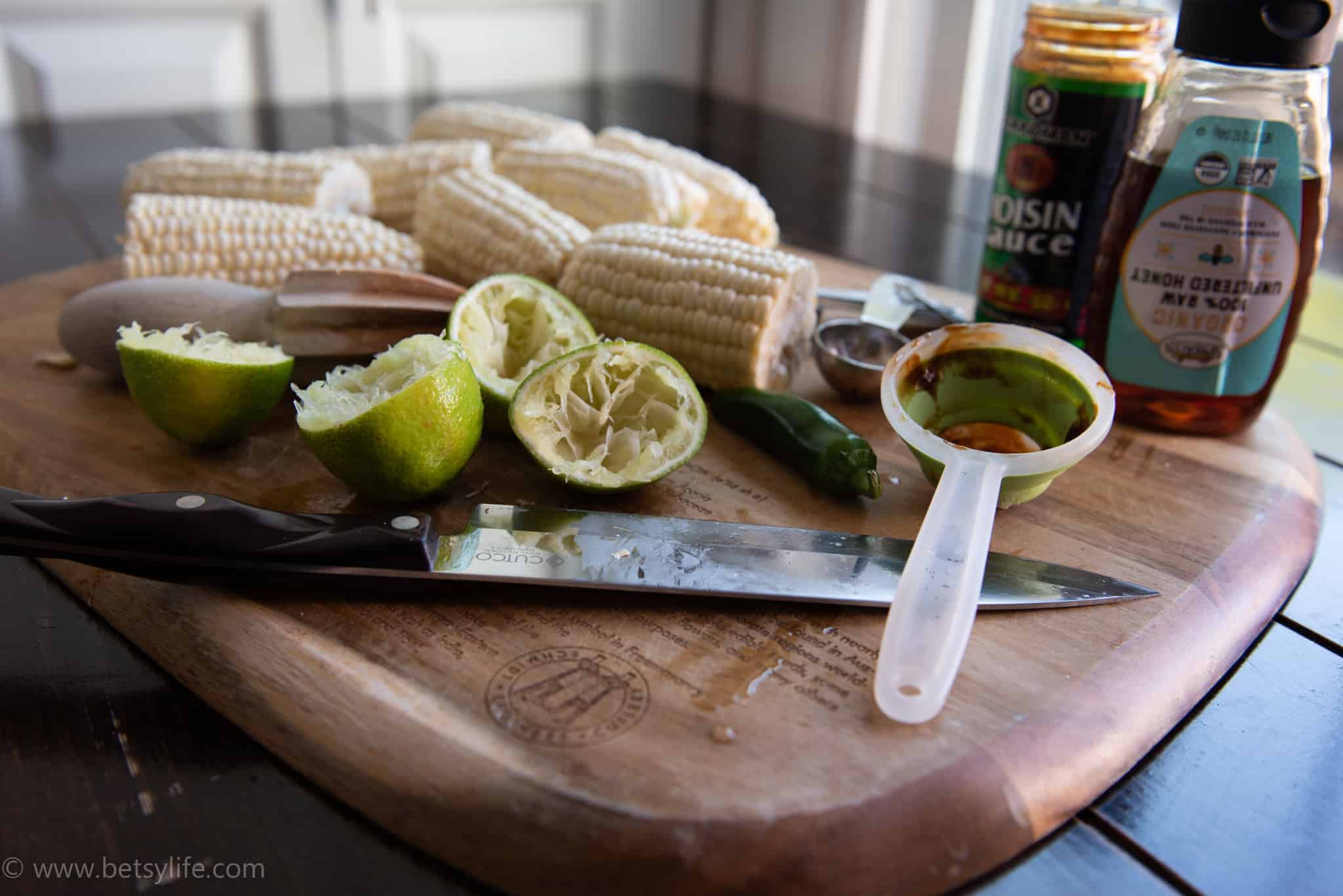 cutting board with cut, juiced limes, hoisin sauce, uncooked corn on the cob, honey, measuring cup and spoons, knife