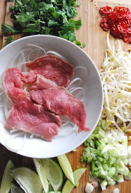 Homemade Pho: slices of beef in white bowl surrounded by cilantro, limes and bean sprouts