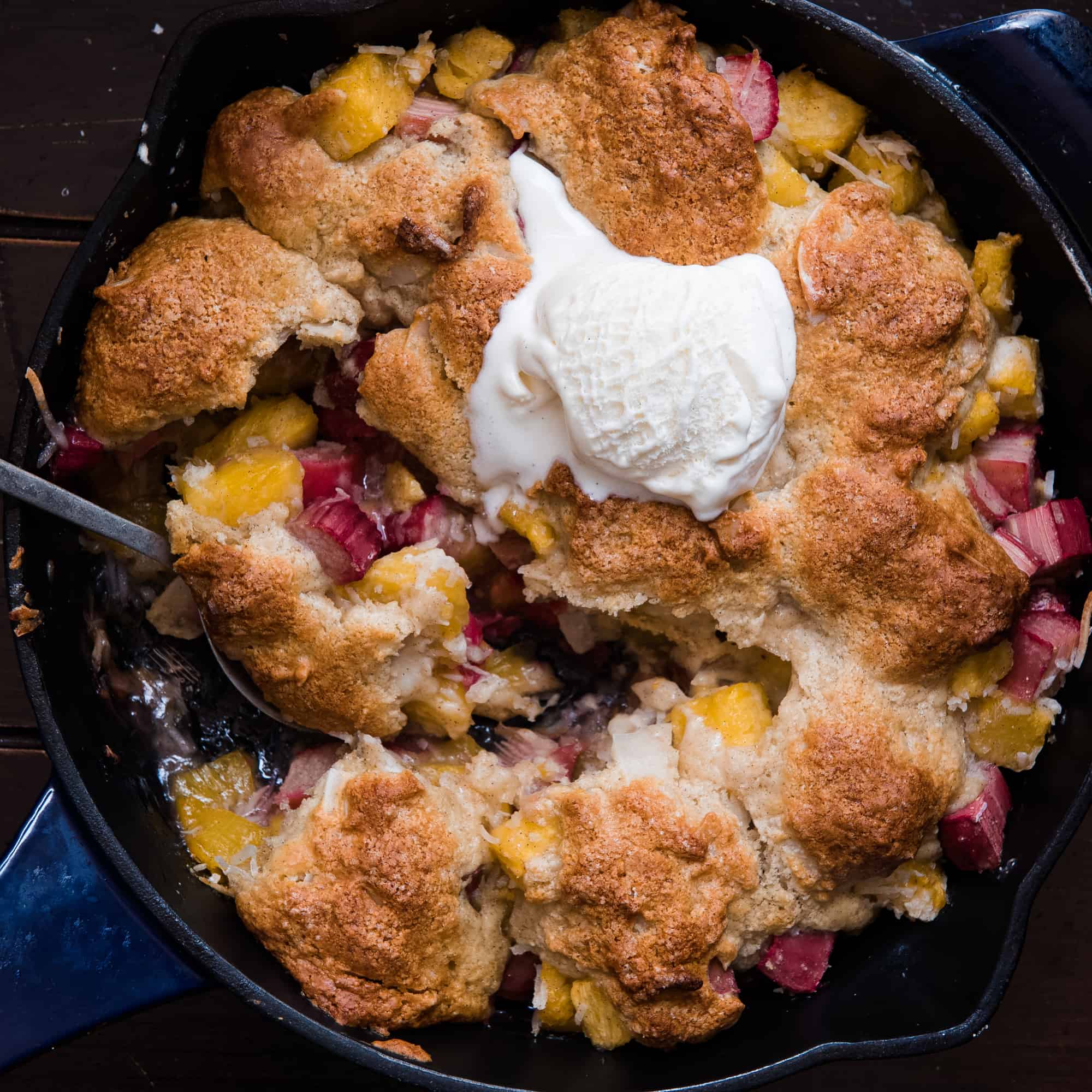 Round pan of cobbler topped with melting vanilla ice cream being scooped out with a spoon 