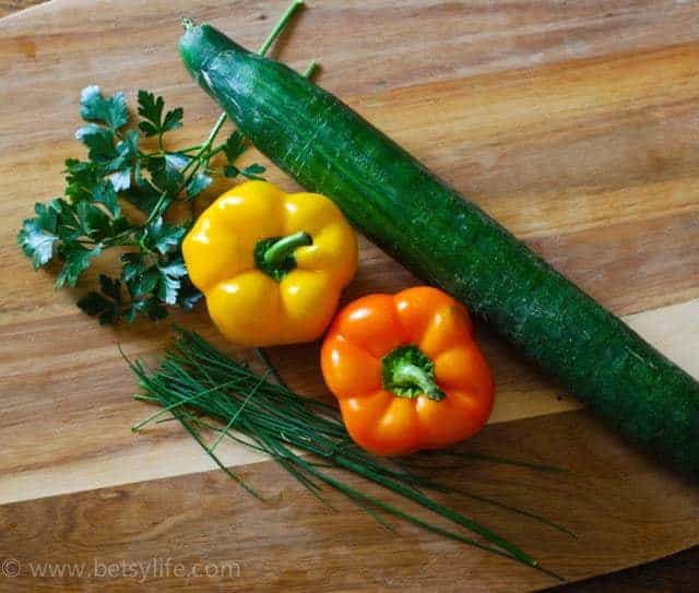 Overhead of cucumber, orange and yellow bell peppers and fresh herbs on a wooden cutting board