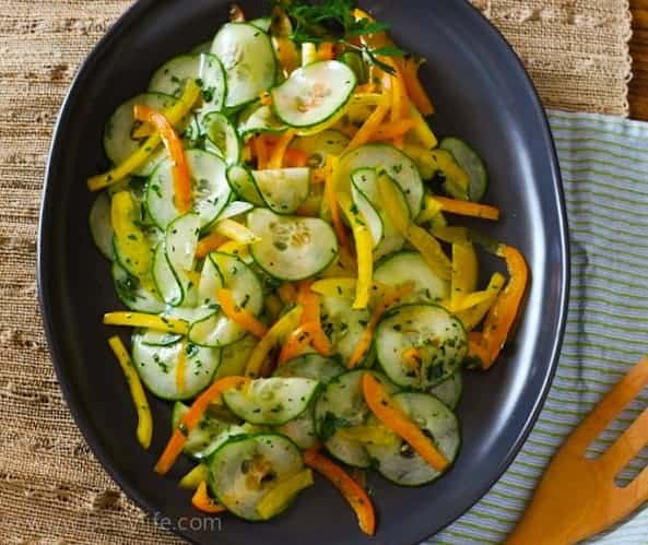 Easy Cucumber Salad. Thinly sliced cucumbers, orange and yellow peppers and fresh herbs on a dark oval serving platter 