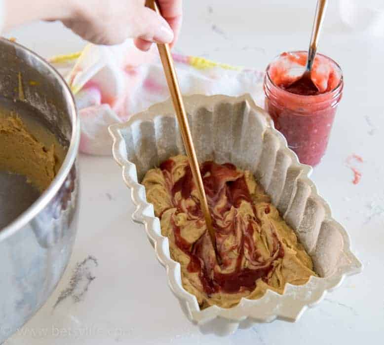 Loaf pan half full of pound cake batter and strawberry glaze. Hand holding a chopstick is swirling the mix together. Metal bowl full of batter on the left and jar of strawberry glaze on the right with a spoon in the jar. 
