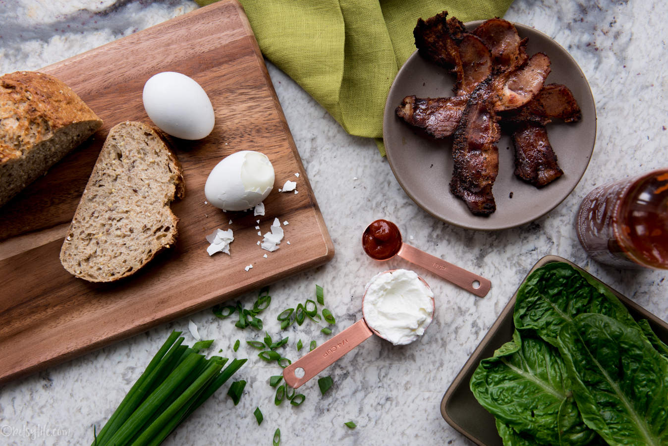Ingredients for sriracha bacon egg salad sandwich on a marble counter. Bread, eggs, bacon, scallions, lettuce, greek yogurt and Sriracha 