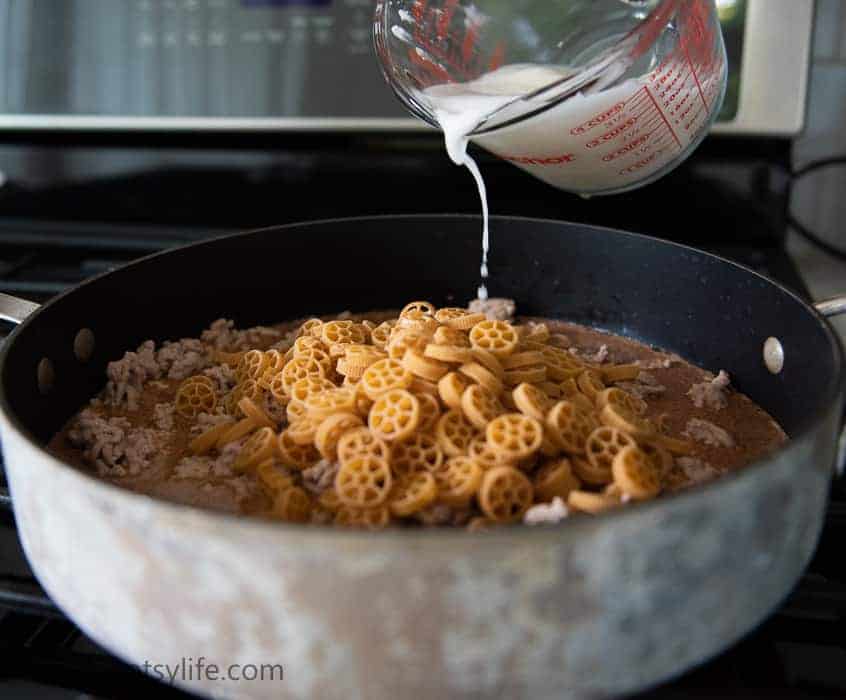 pan of dried pasta and ground turkey with white liquid being poured over the top 