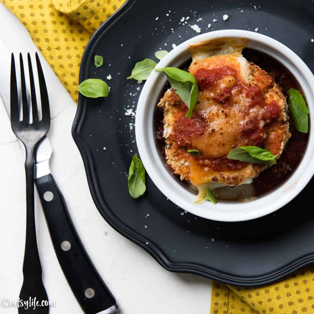Ramekin with a single serving of eggplant parmesan on a black plate with a yellow napkin