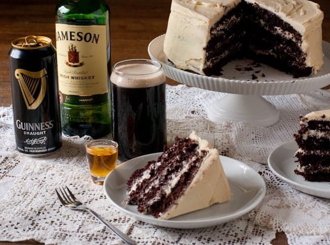 Boozy Chocolate Stout Cake on a white cake stand with two slices removed and on plates next to guinness and jameson bottles 