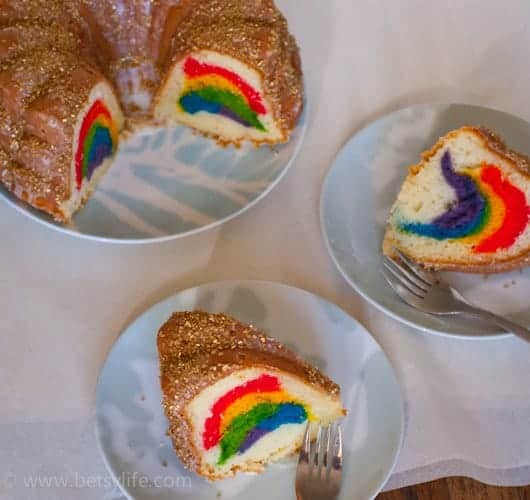 Overhead of a bundt cake on a platter with two slices removed. Slices are on small plates on either side with forks. Cake and slices are golden brown and topped with gold sprinkles on the outside and the inside shows rainbow colored layers of cake. 