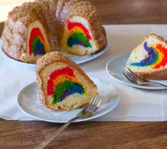 Two slices of white bundt cake on small light blue plates. Fully cake on a platter in the background. All slices reveal layers of rainbow colors on the inside. 