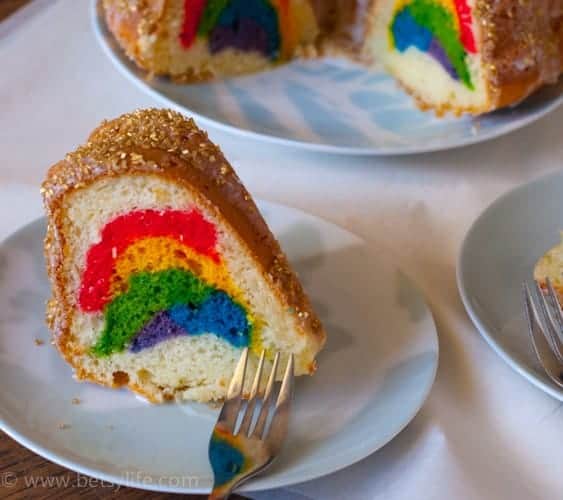 Slice of a white bundt cake with a rainbow motif on the inside. Layers of red, yellow, green, blue and purple cake. Topped with gold sprinkles on a blue plate with a fork. 