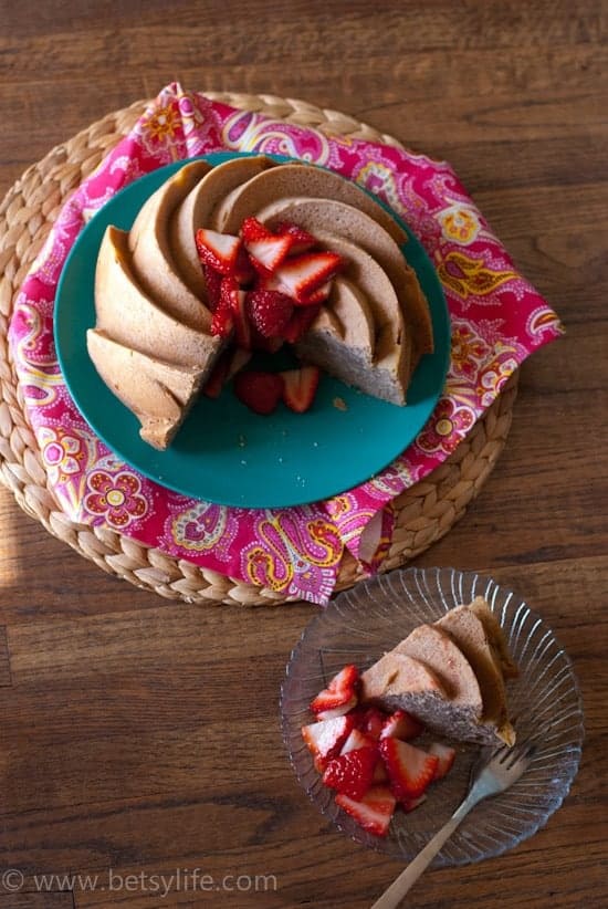Strawberry Rhubarb Bundt Cake overhead on a wooden background. Cake is sitting on a teal plate on top of a pink printed napkin. Slice removed and is plated on a glass plate with a fork resting on the plate. 