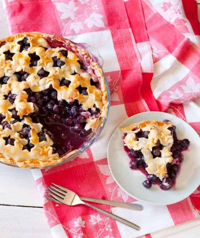 Blueberry pie with star shaped cut out crust on a red and white checked table cloth. Slice removed and on a plate to the right. Forks in lower portion of the photo