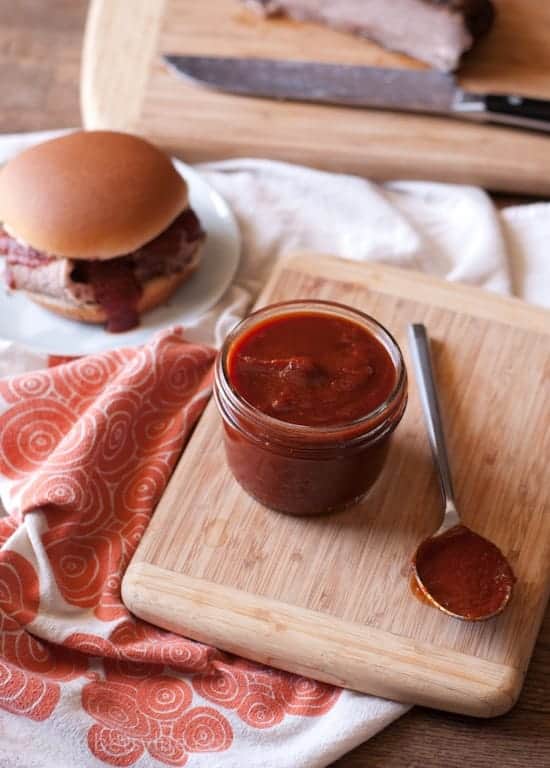 easy bbq sauce recipe. jar of homemade bbq sauce in a jar with a spoon on the side. Pulled pork sandwich with oozing bbq sauce out of focus in the background 