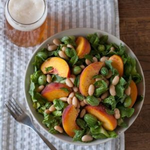Overhead photo of White Bean and Peach Salad with a Lemon-Mint Vinaigrette in a bowl on a gray and white striped towel with a full glass of beer in the upper left corner and a fork in the lower left corner