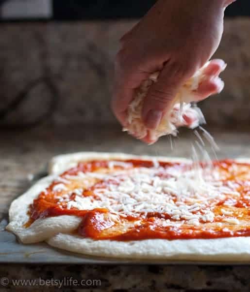 Mozzarella cheese being sprinkled over a pizza dough round topped with marinara sauce 