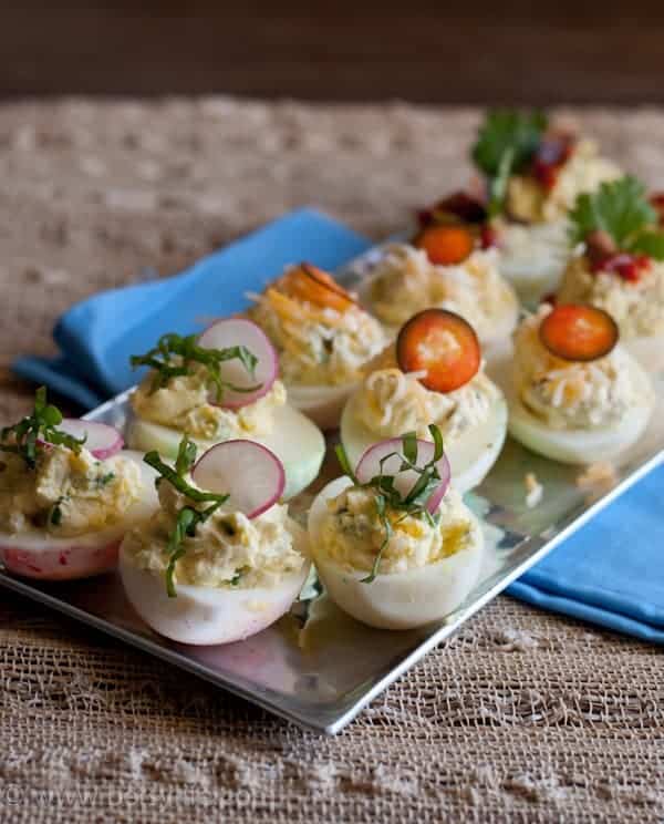 Silver tray of deviled eggs topped with herbs, radishes and jalapenos on a blue napkin and a natural woven mat