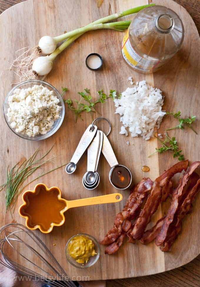 Ingredients for bacon potato salad with blue cheese. Bowl of blue cheese crumbles, spring onions, chives, parsley, bacon, mustard, apple cider vinegar and olive oil. Whisk and measuring spoons. 