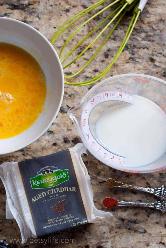 overhead photo of a bowl of whisked eggs, a whisk a measuring cup full of milk and a blog of Kerry Gold aged cheddar cheese 