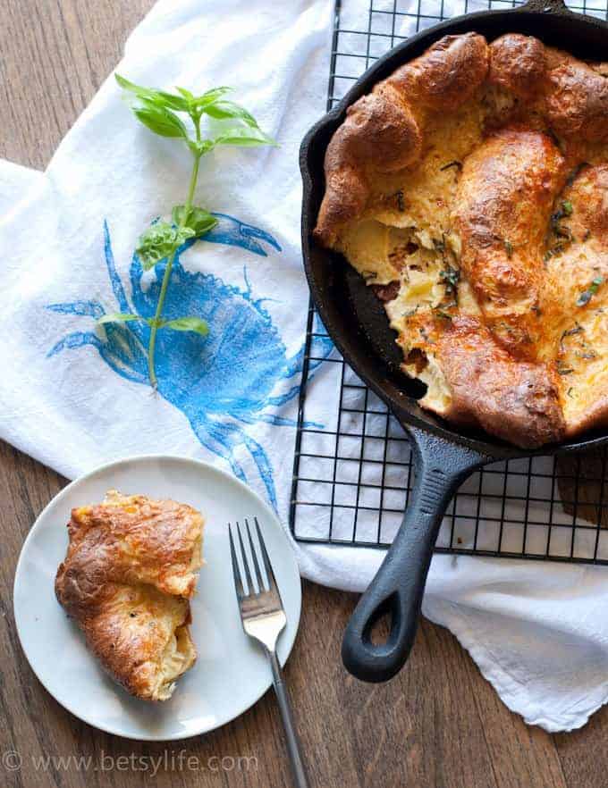 Cheddar dutch baby pancake in a cast iron skillet with a serving torn from it. Serving on a small plate next to skillet with a fork. Skillet sitting on a cooling rack with a white towel underneath. Stem of fresh basil on the side. 