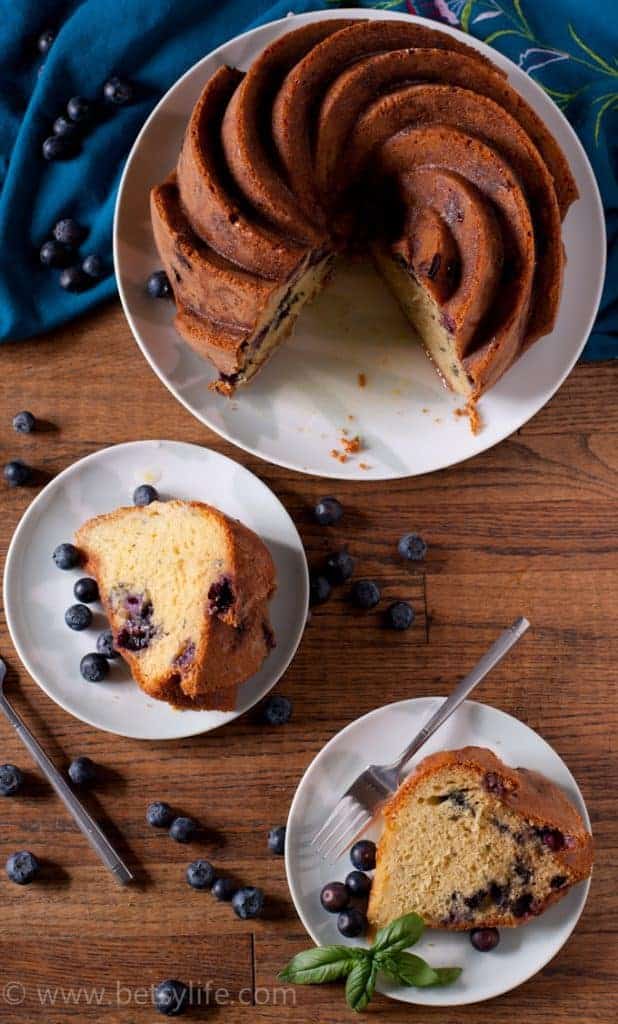 overhead photo of basil blueberry bundt cake with two slices cut out and on separate white plates. Blueberries scattered around