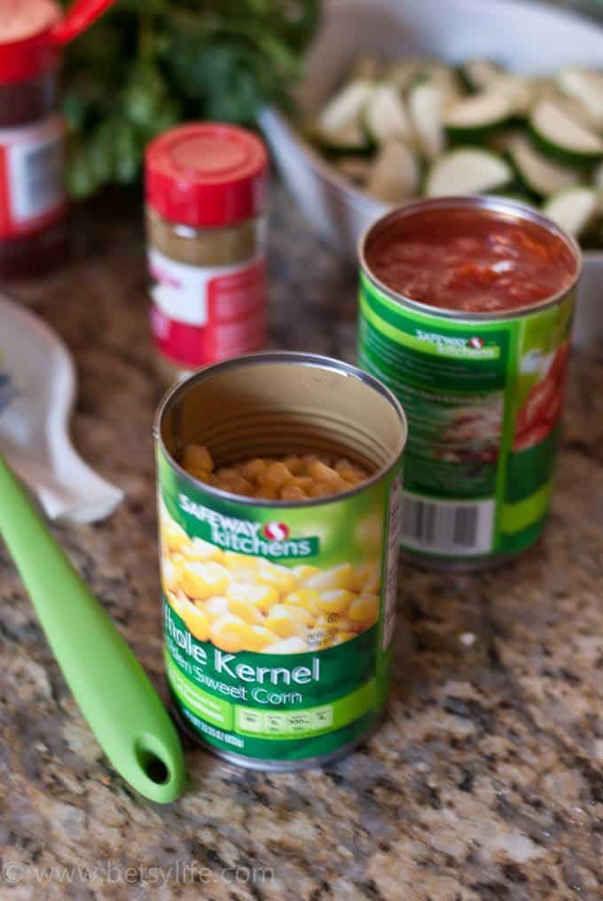 Open can of corn and diced tomatoes on a counter next to a green spatula. Sliced zucchini and a spice bottle out of focus in the background 