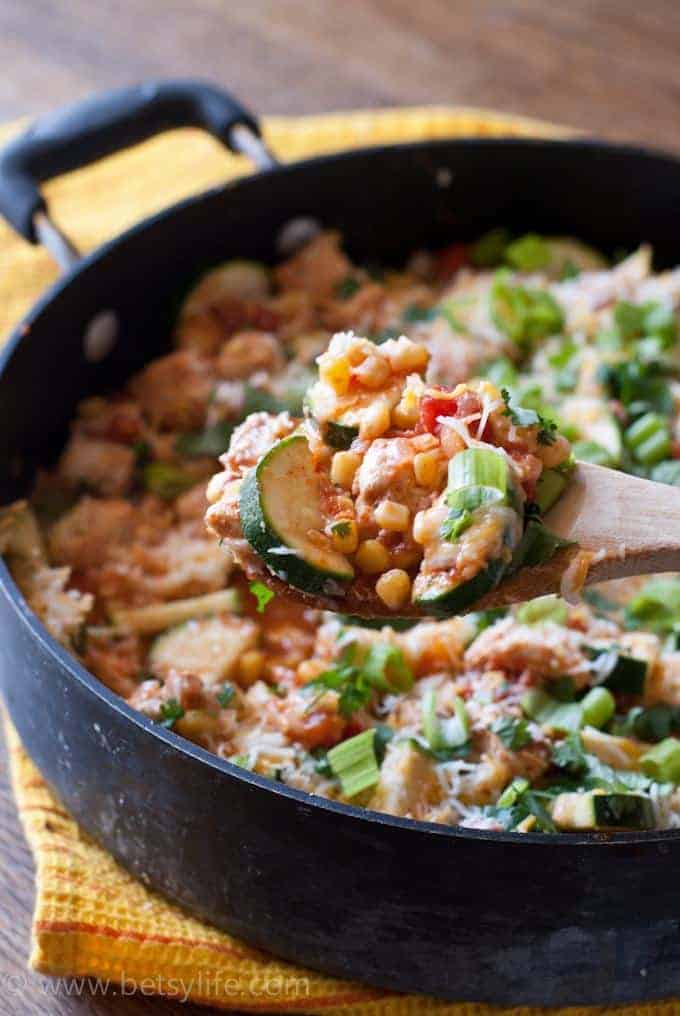 Close up of a wooden spoon filled with corn, chicken and zucchini in front of a large skillet filled with the same mixture
