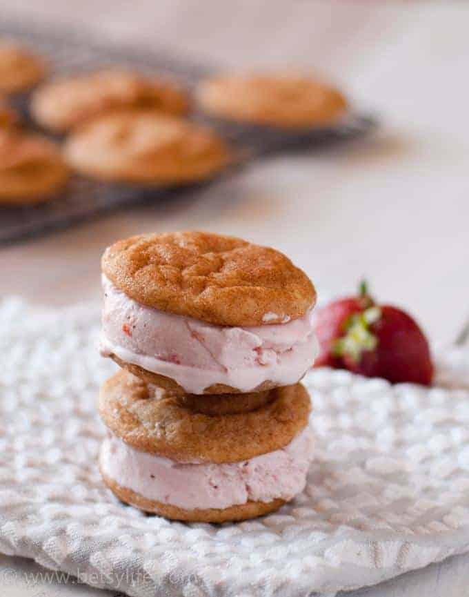 Two Snickerdoodle and Roasted Strawberry Ice Cream Sandwiches stacked with a cooling rack filled with snickerdoodle cookies out of focus in the background