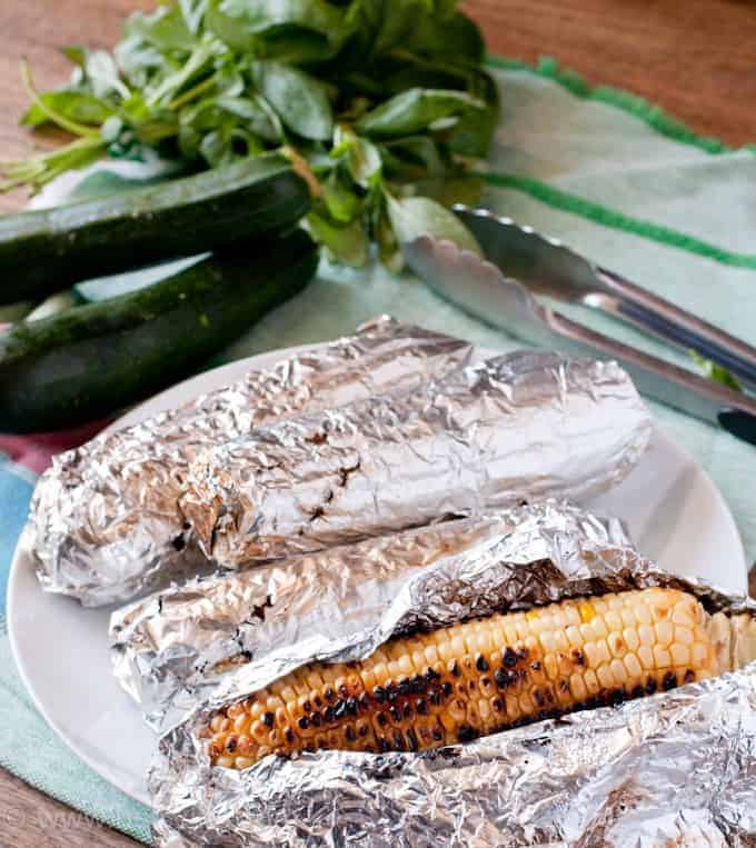 Grilled corn in foil with the foil being pulled back. Zucchini and fresh herbs in the background