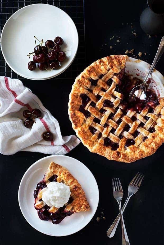 Overhead of cherry pie. One slice removed on a white plate. Another white plate of fresh cherries. White linen draped over a metal cooling rack. All on a dark background