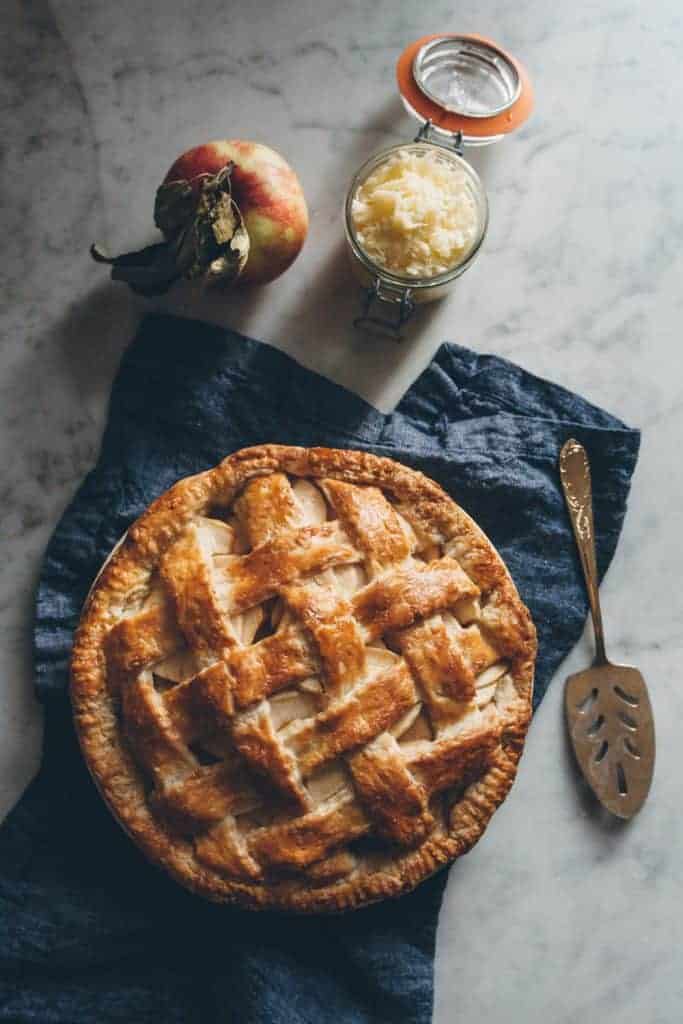 Overhead photo of apple pie with a lattice top on a navy blue napkin. Latch lid jar of shredded cheddar cheese and a whole apple with a leaf attached on the side. 