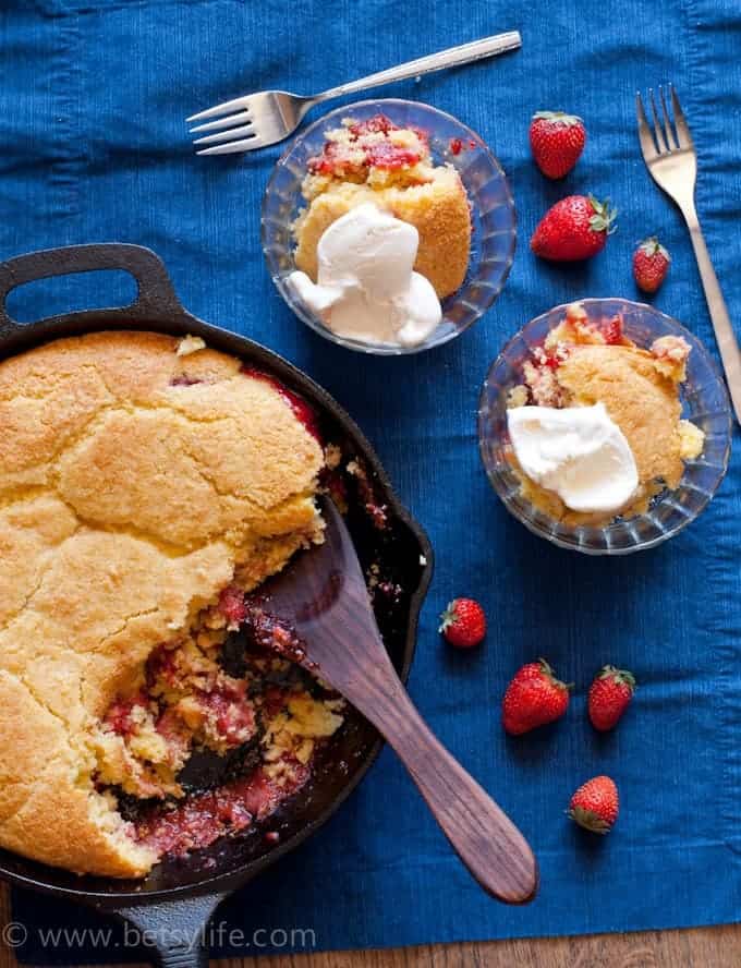 Dark blue background with a cast iron skillet filled with strawberry cobbler with cornbread topping. Wooden spoon in the cobbler and two servings are spooned out into glass bowls on the side. Two forks and 7 whole strawberries scattered around. 