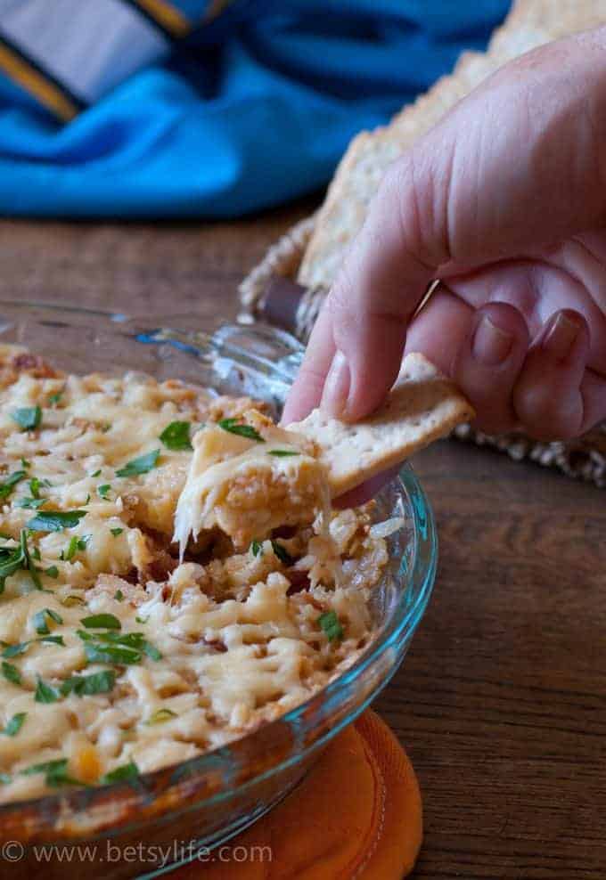 Hand dipping a cracker into a round glass baking dish of Cheesy Tater Tot Bacon Dip 