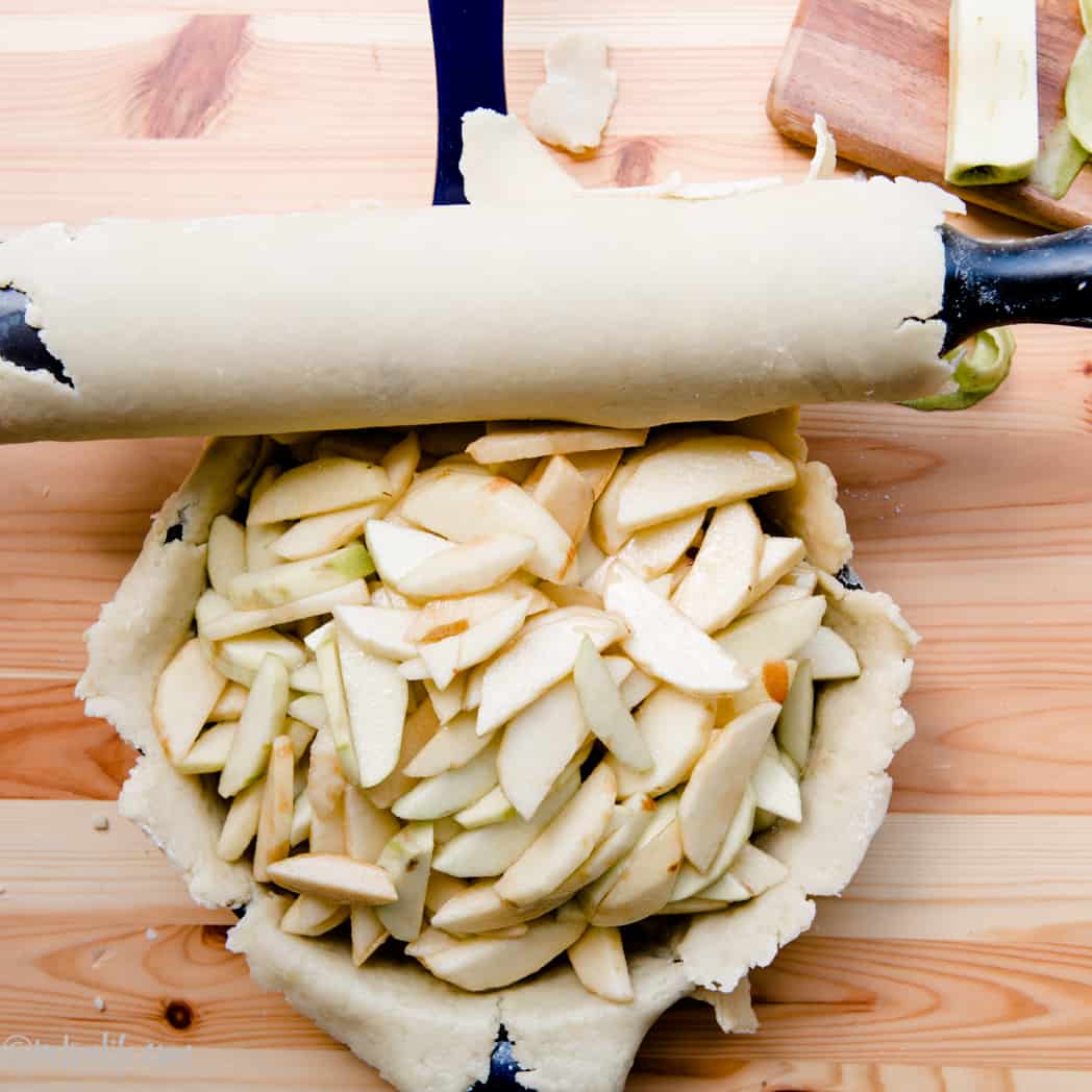 Top pie crust being rolled over a pie filled with apple slices ready to be baked