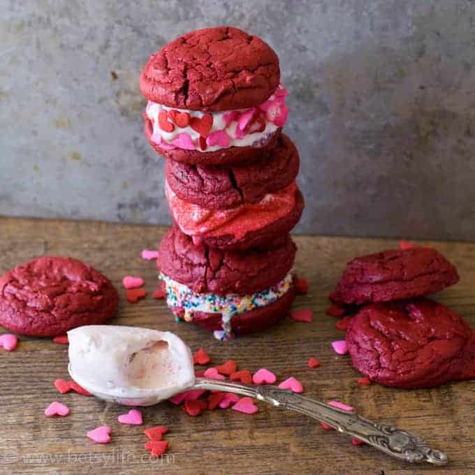 three red velvet ice cream sandwiches stacked with three loose cookies, sprinkles and a scoop of ice cream surrounding stack