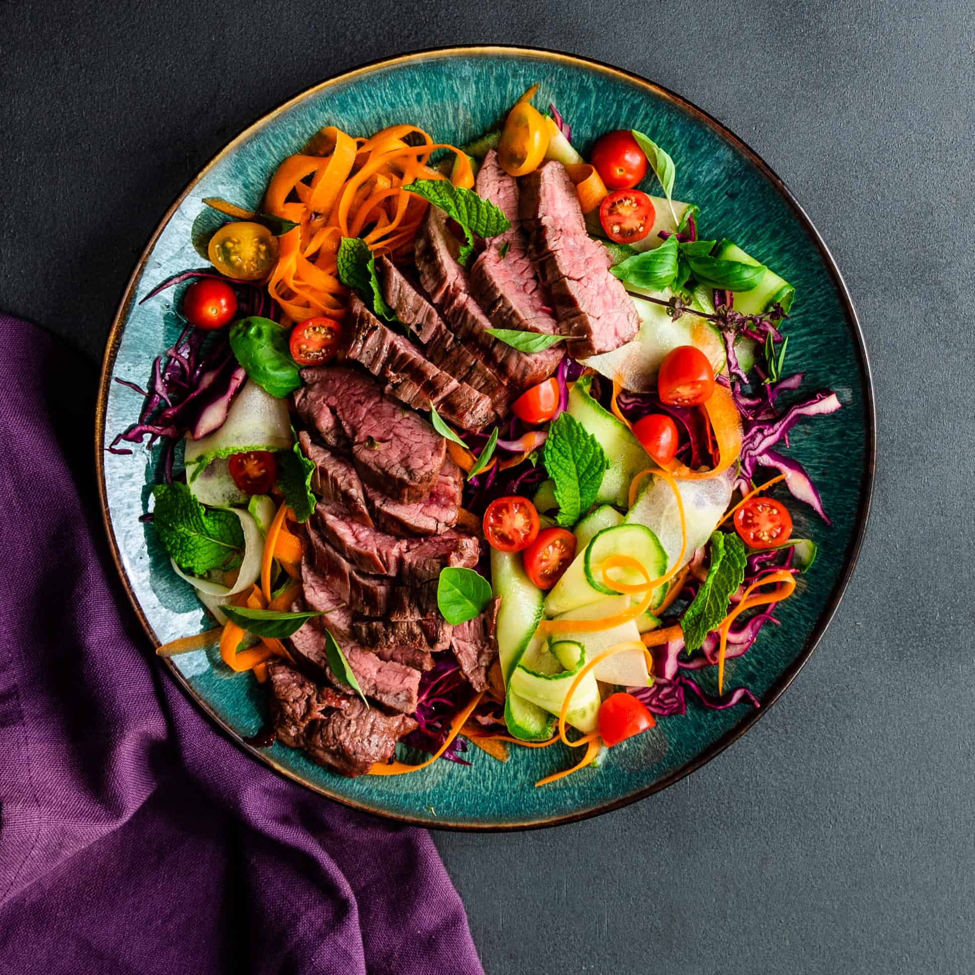 Blue plate with grilled steak salad with vegetable noodles and a purple napkin