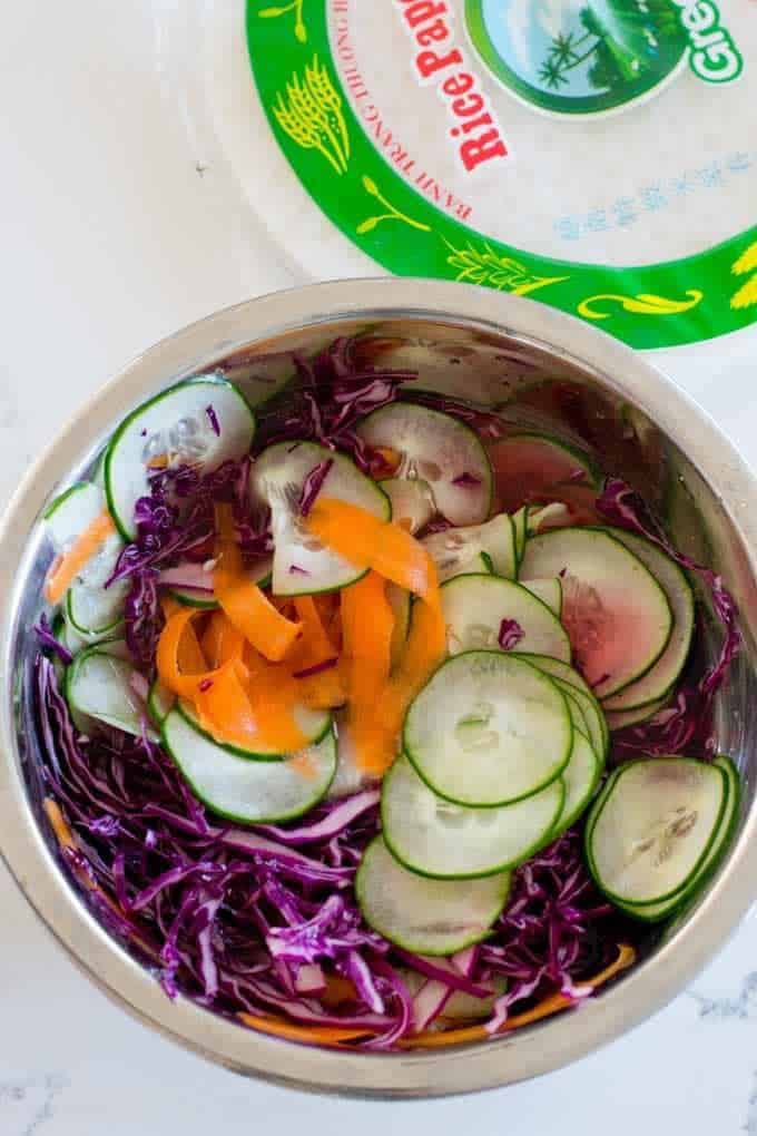 Bowl filled with thinly sliced cucumber, carrots and cabbage. Package of spring roll wrappers on the side
