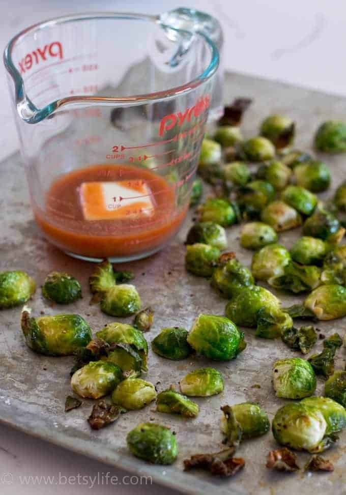 roasted brussels sprouts on a sheet pan next to a measuring cup with buffalo sauce inside 
