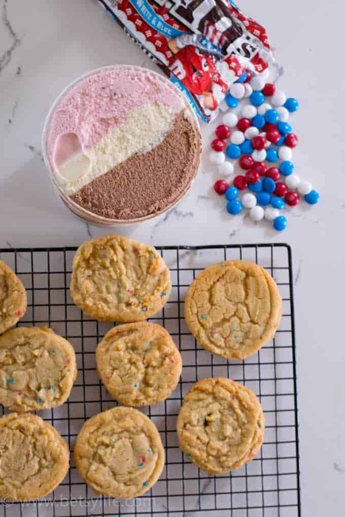 Overhead of funfetti cake mix cookies cooling on a wire rack next to a round container of neapolitan ice cream and a spilled bag of red, white and blue m&ms