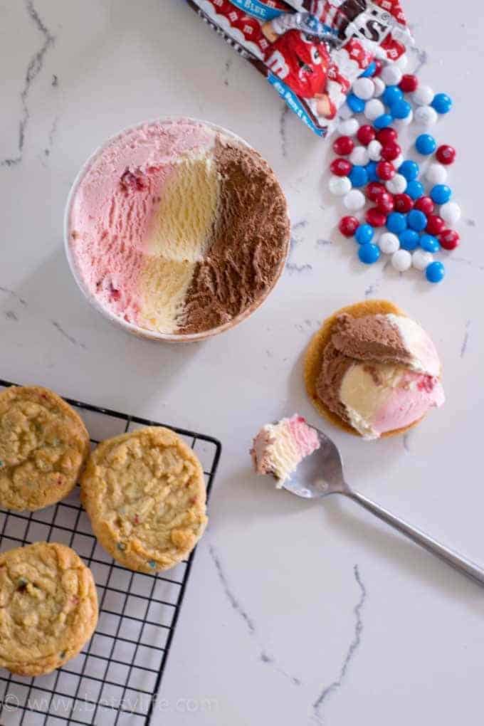 Funfetti cake mix cookies on a wire rack next to a round container of neapolitan ice cream with a scoop removed and on a cookie on the counter. Spilled bag of red, white and blue m&ms