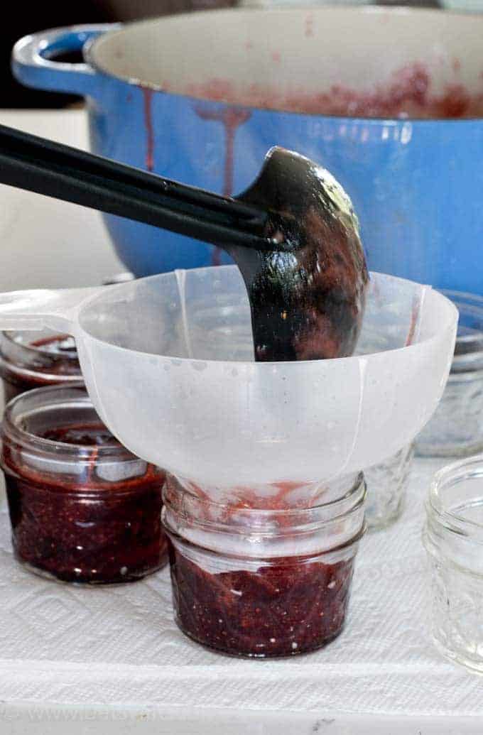 homemade strawberry jam being ladled into glass jars with a black ladle. Blue dutch oven with jam drips on the edge out of focus in the background 