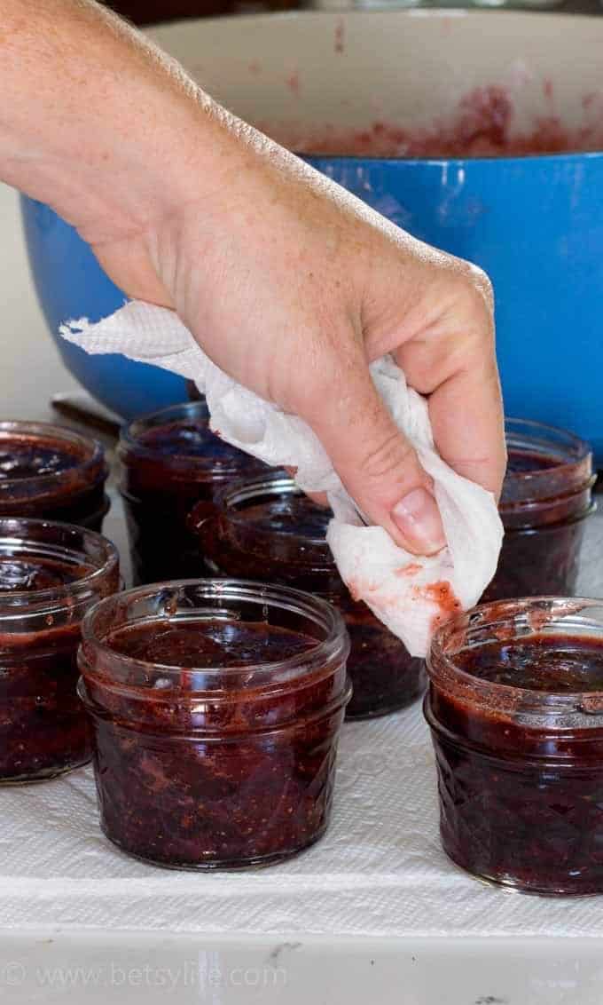 hand with damp paper towel wiping the rim of 7 clear glass jars lined up, filled with homemade strawberry jam