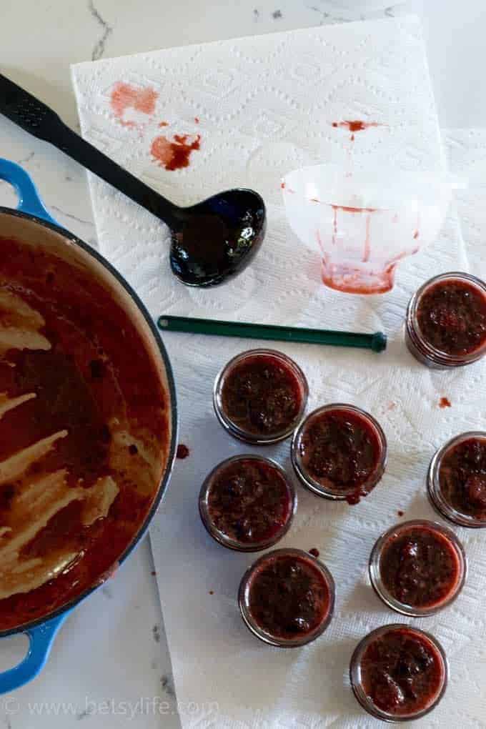 overhead of a white counter top lined with paper towel covered in 8 jam jars filled with homemade strawberry jam. A dutch oven filled with jam and a black ladle to the left 