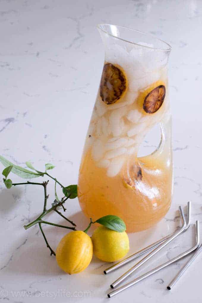 pitcher of best lemonade recipe with ice and grilled lemon slices floating in it. Two fresh lemons and 4 metal straws scattered around the pitcher