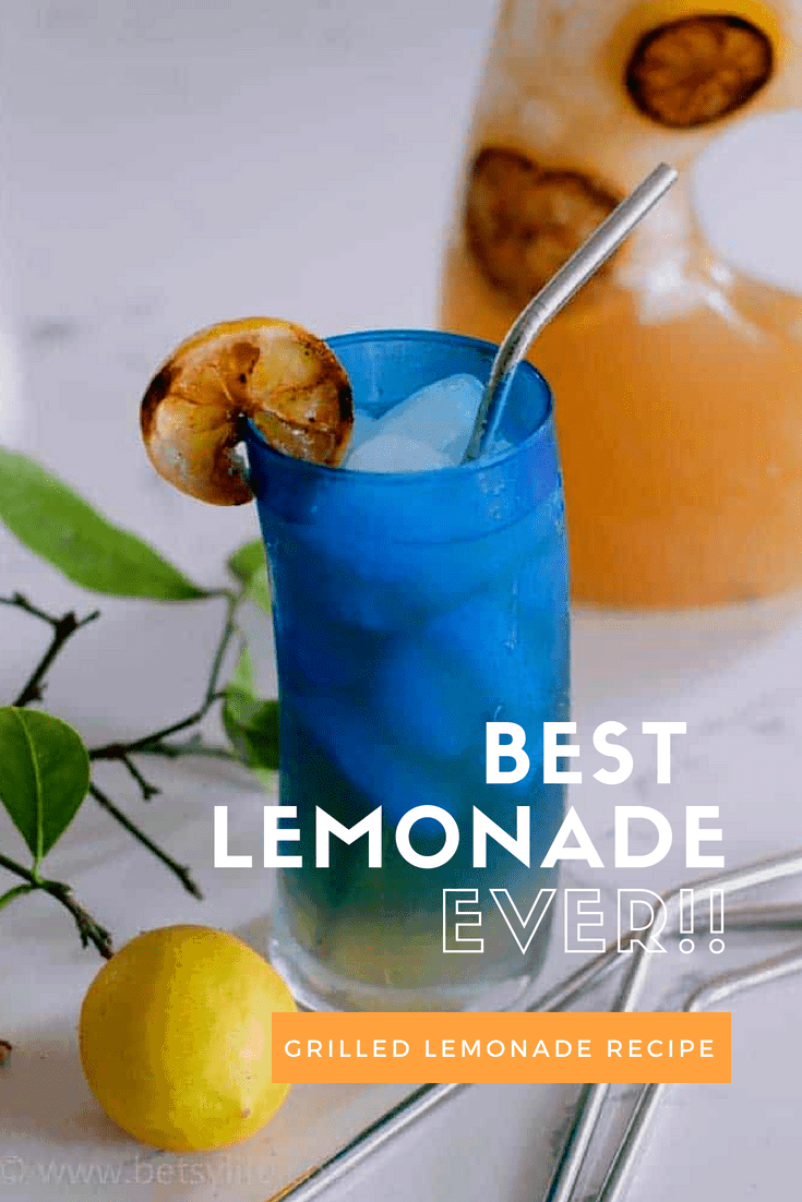 close up of the best lemonade recipe in a blue glass with a grilled lemon slice on the edge of the glass. clear pitcher out of focus in the background. One fresh lemon and 3 metal straws scattered around in front