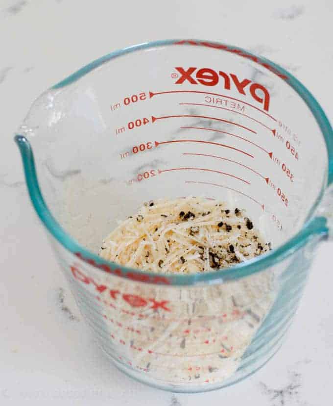 Measuring cup filled with shredded parmesan cheese and fresh cracked pepper 