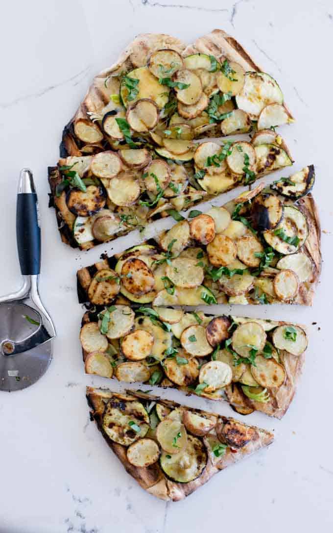 Grilled Potato Pizza overhead photo. Sliced on a white marble surface with a pizza cutter on the left side 