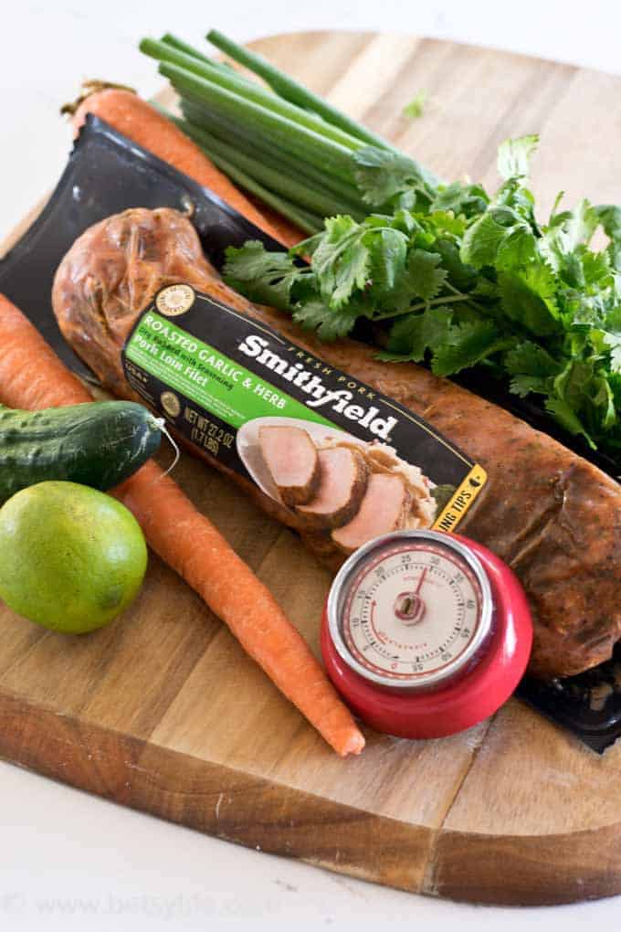 ingredients for a healthy 30 minute meal on a cutting board. Marinated pork tenderloin, carrot, cucumber, lime, cilantro, green onion