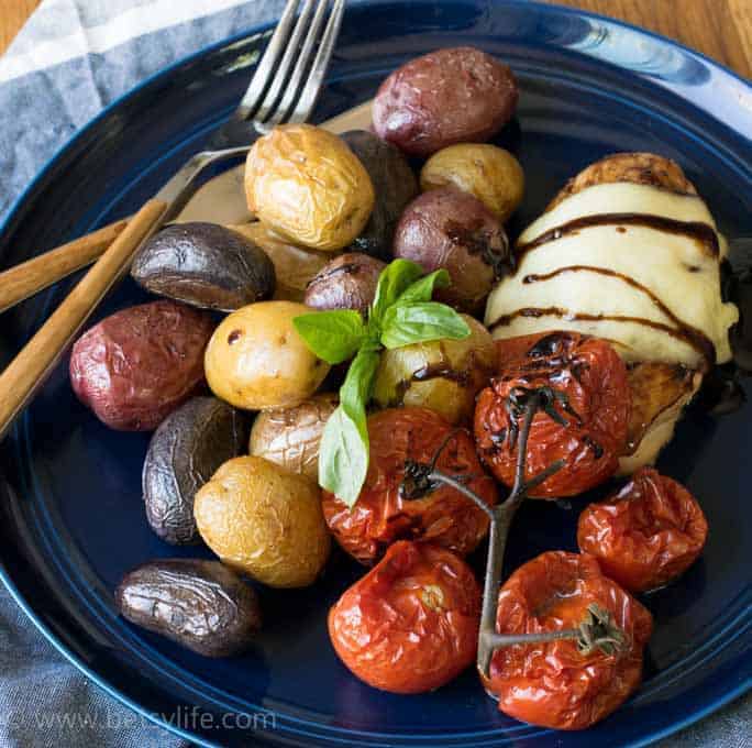 blue plate on a blue and white napkin. Chicken breast topped with melted cheese, roasted rainbow baby potatoes and roasted tomatoes. Fork and knife with wooden handles. 