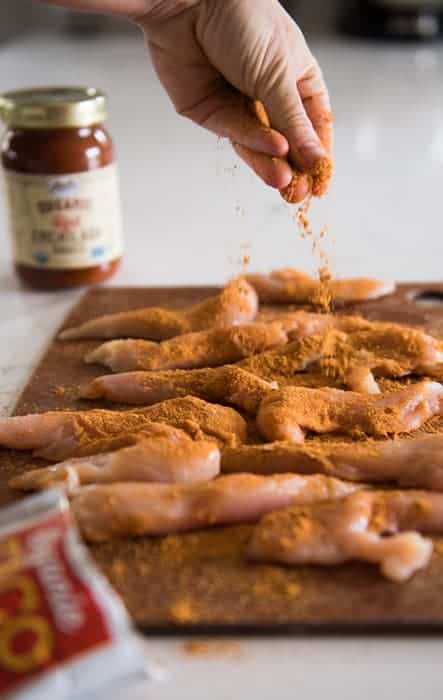 Hands sprinkling taco seasoning over chicken tenders on a wood cutting board. Jar of red enchilada sauce out of focus in the background