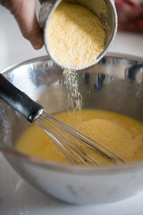 cornmeal pouring from a measuring cup into a silver bowl with a whisk
